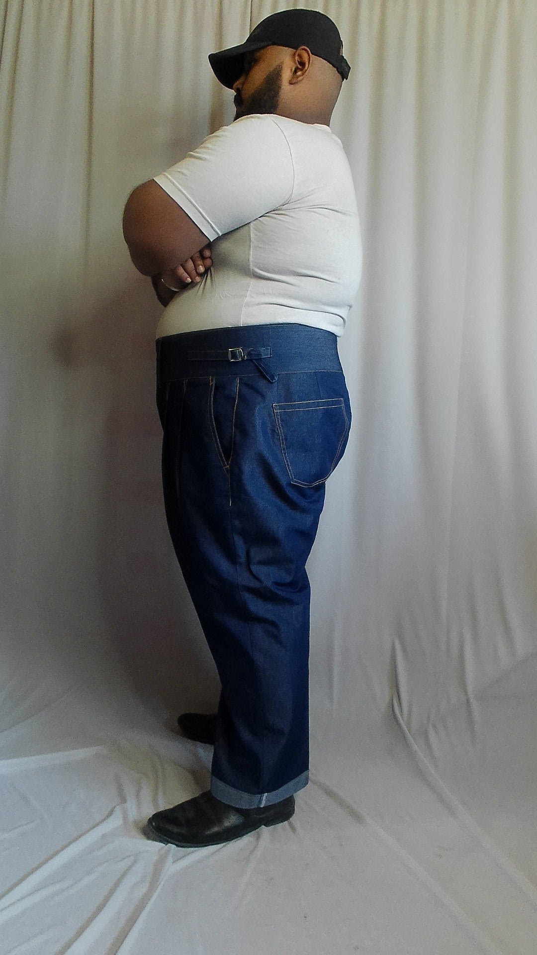 Man on a white surface wearing a white shirt, blue gurkha jeans trouser, and black shoes.